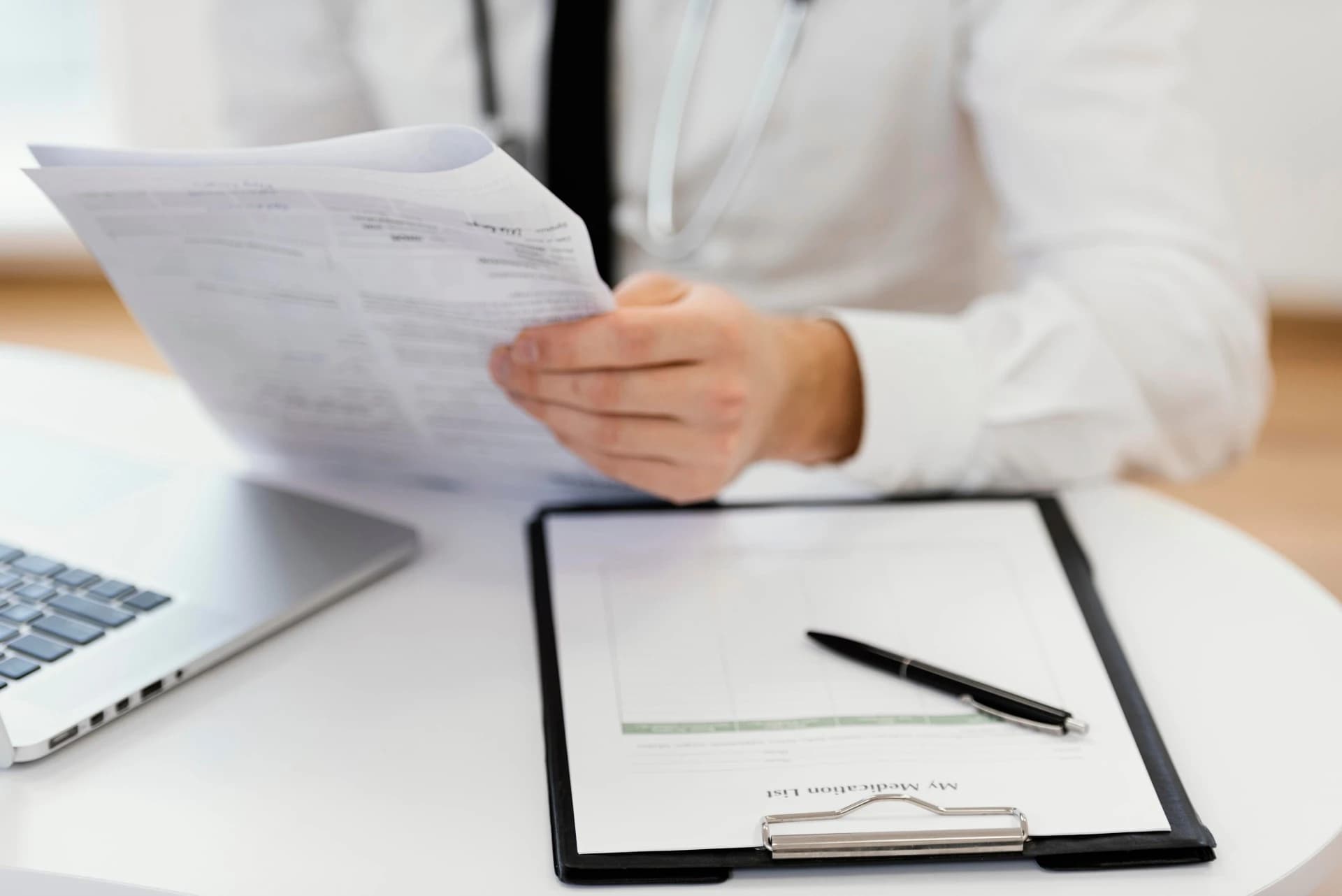 doctor-sitting-on-desk-holding-medical-records-with-laptop-and-form-placed-on-desk